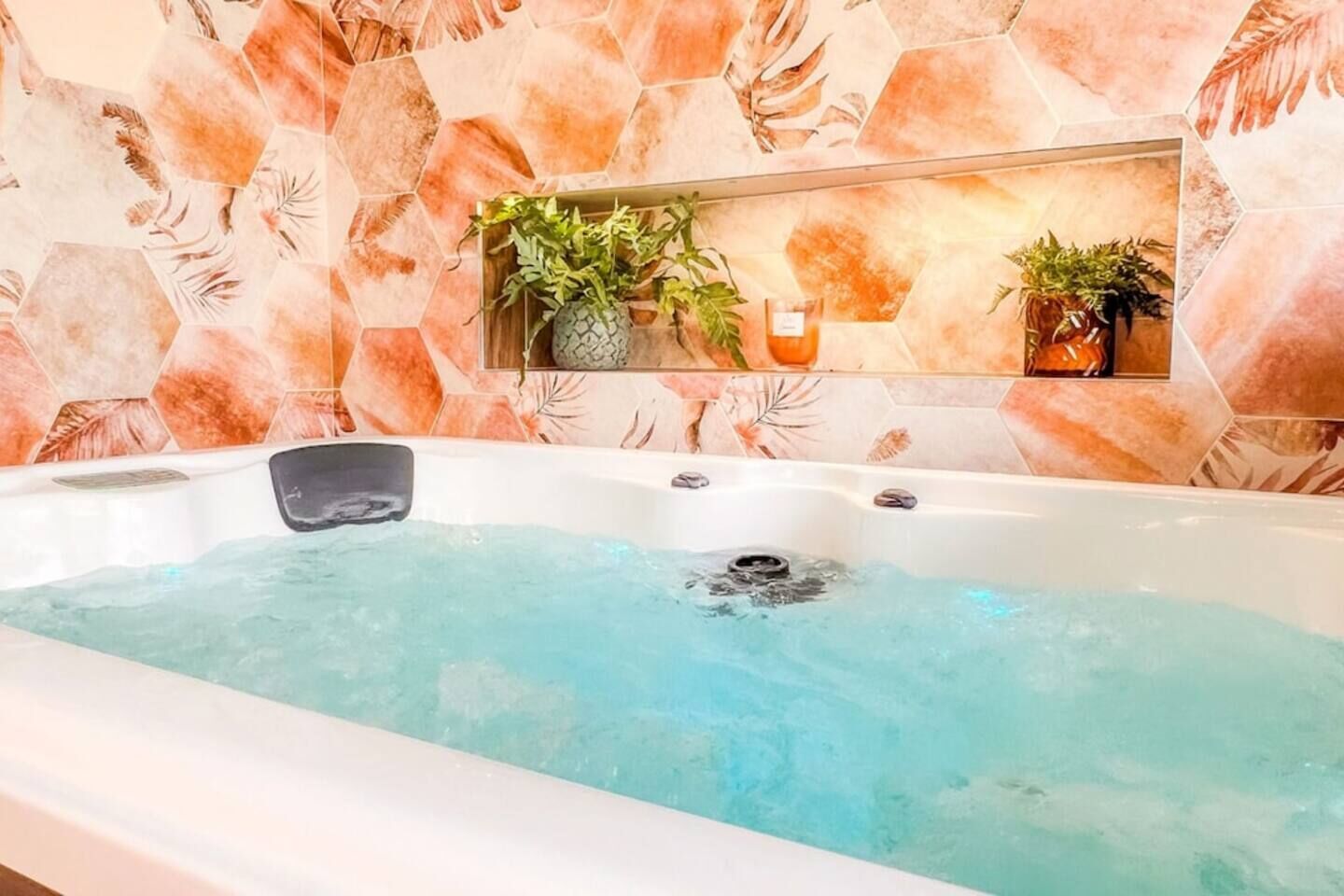 Uma banheira de hidromassagem com parede de azulejos hexagonais, um prateleira com plantas e uma vela.