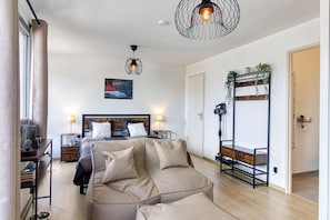 A modern bedroom with a bed, bedside tables, a hanging light fixture, a painting, a plant shelf, and a sofa. - Le Studio des Peupliers - 30m de la plage (Bray-Dunes)