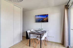 A dining area with a table set for four, a flat-screen TV mounted on the wall, and a sliding door on the left. - Le Studio des Peupliers - 30m de la plage (Bray-Dunes)