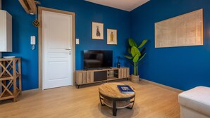 A room with a blue wall, a white door, a television, a wooden cabinet, a potted plant, and a small round table with a book. - Vigie d'Opale - Nausicaá & plage à 700 m (Boulogne-Sur-Mer)