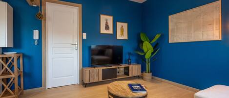 Een kamer met een blauwe wand, een witte deur, een televisie, een houten kast, een potplant en een kleine ronde tafel met een boek.