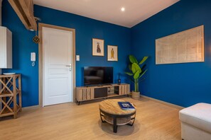 Ein Zimmer mit blauer Wand, weißer Tür, Fernseher, Holzschrank, Blumentopf und rundem Tisch mit einem Buch.