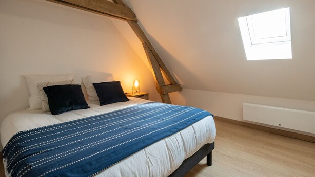 A bedroom with a sloped ceiling, a bed with white bedding and a blue striped blanket, two black pillows, a nightstand with a lamp, and a window with blinds. - Vigie d'Opale - Nausicaá & plage à 700 m (Boulogne-Sur-Mer)