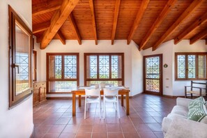 Ein geräumiger Raum mit hoher Holzbalkendecke, großen Fenstern mit Kreuzverstrebungen, einem Esstisch mit Stühlen und einem weißen Sofa.