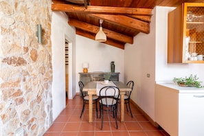 A dining area with a stone wall, wooden ceiling beams, a table with chairs, and a kitchen area with cabinets and a plant. - VILLA ALICE with sea view in Torre delle Stelle (TORRE DELLE STELLE)