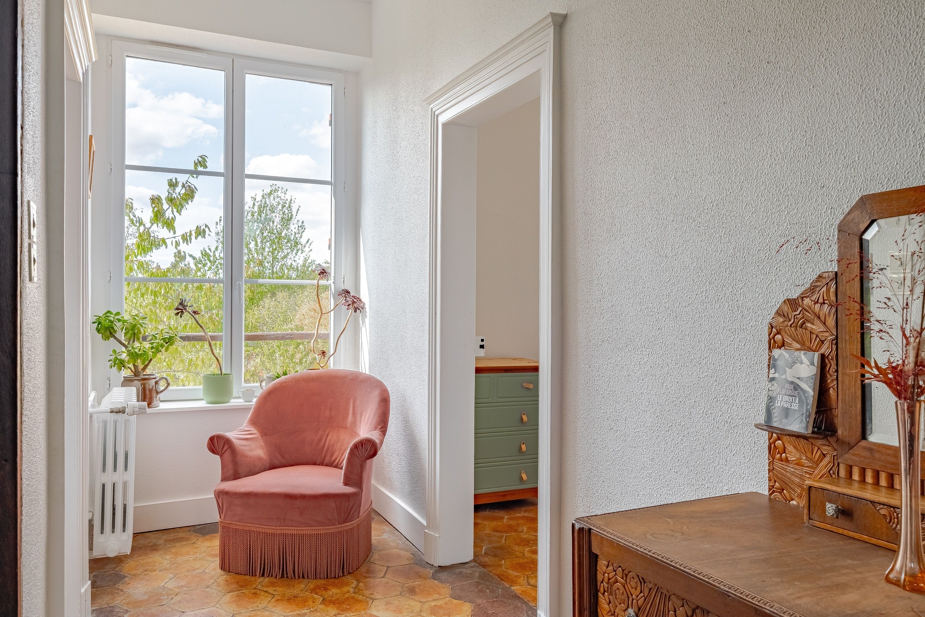 Une pièce avec une grande fenêtre, un fauteuil en osier, une commode en bois et un radiateur.