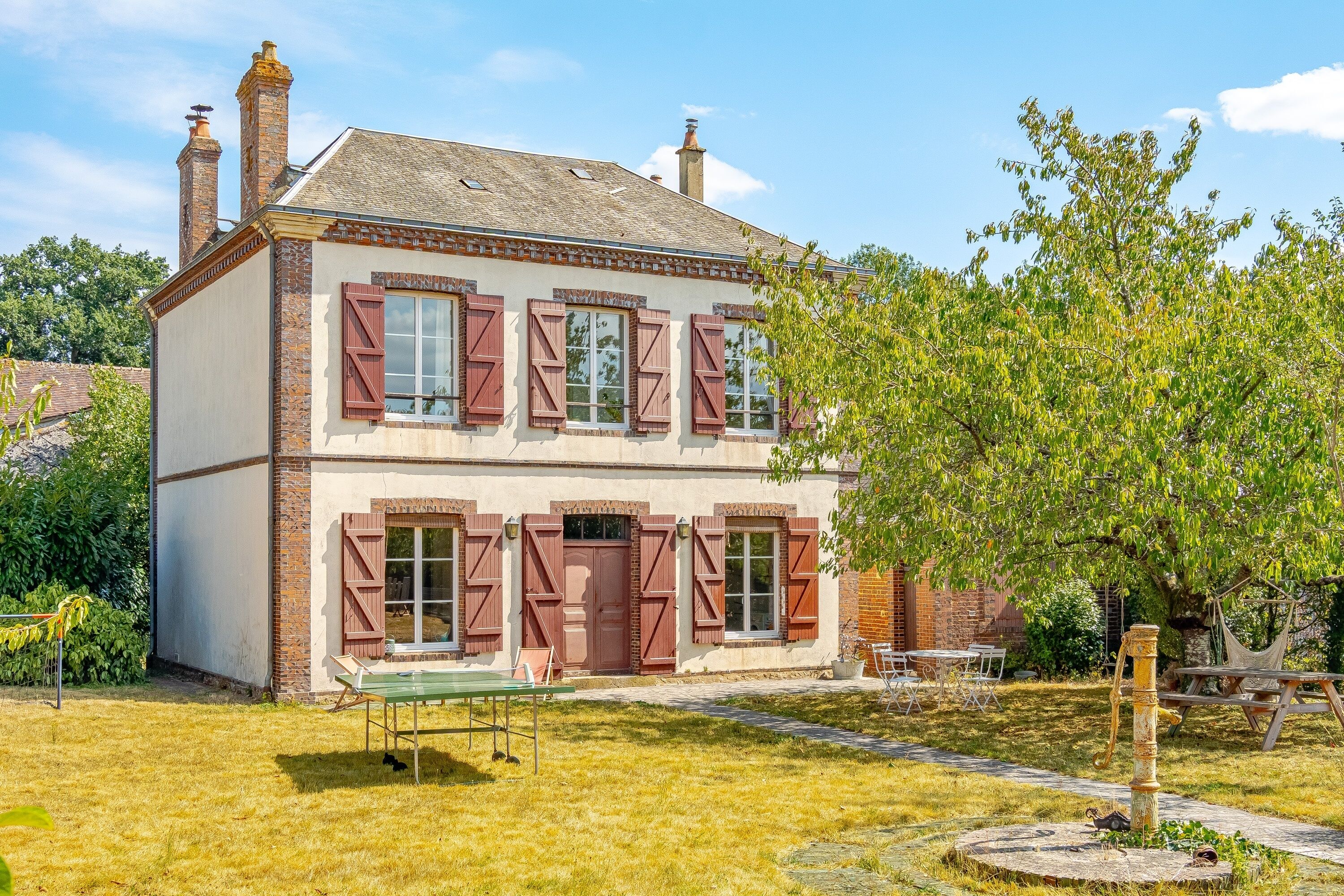 Une maison à deux étages avec une cheminée en briques, des volets et un jardin agrémenté d’une table et de chaises.
