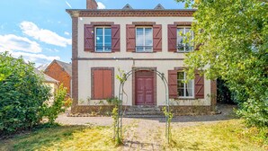 Une maison à deux étages dotée d’une façade symétrique, avec une entrée centrale surmontée d’un arc décoratif, flanquée de fenêtres avec volets et agrémentée d’une cheminée en briques.