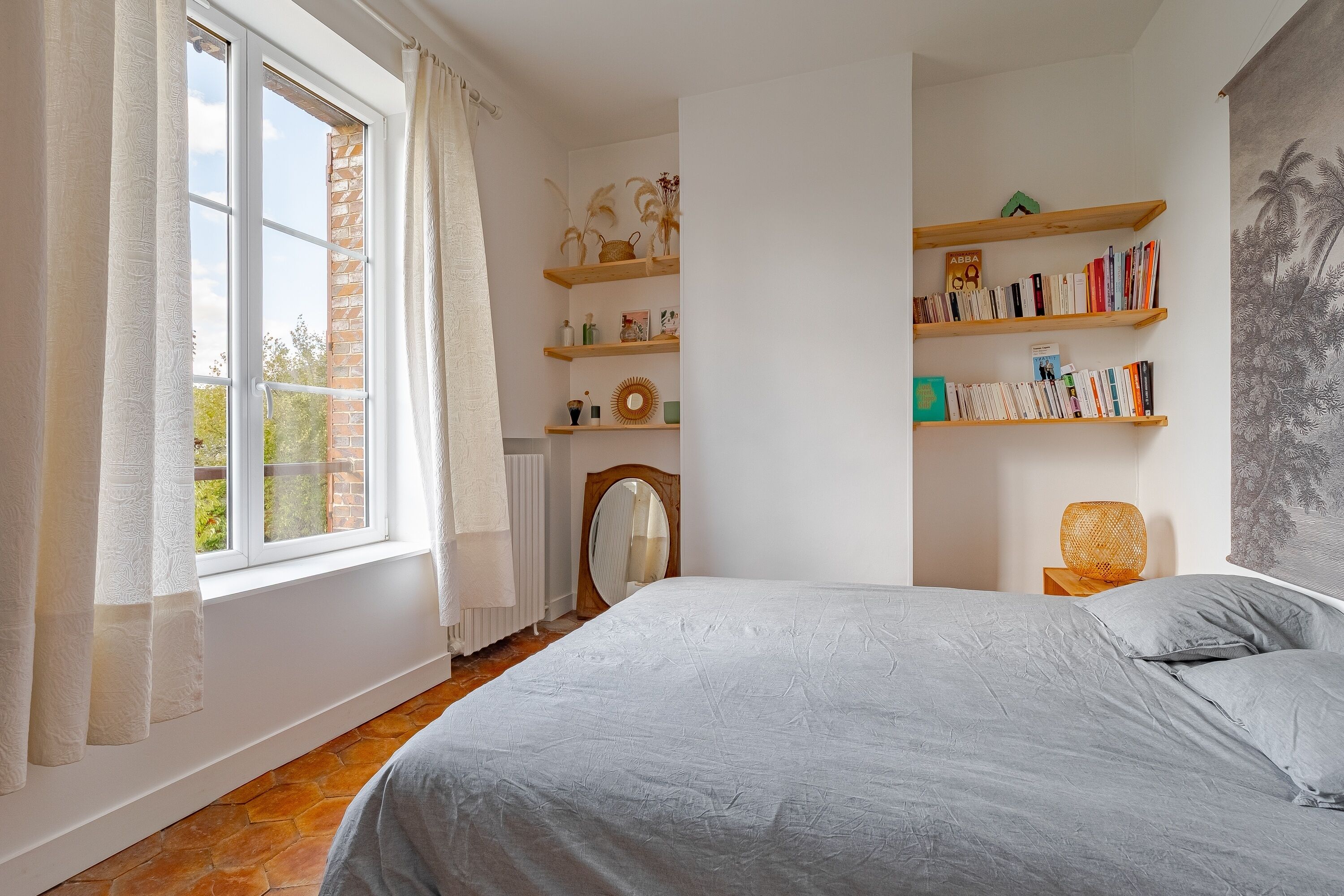 A bedroom with a bed, a window with curtains, a radiator, a bookshelf, and a wall with a painting.