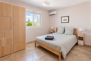 Un dormitorio con cama, armario, ventana, aire acondicionado, mesita de noche, lámpara y un cuadro enmarcado en la pared.