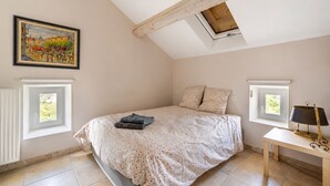 A bedroom with a sloped ceiling, a bed with a floral bedspread, two pillows, a bedside table with a lamp, and a painting on the wall. - La Maison Rose - Avec Piscine Privée (Barjac)
