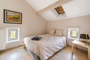A bedroom with a sloped ceiling, a bed with a floral bedspread, two pillows, a bedside table with a lamp, and a painting on the wall. - La Maison Rose - Avec Piscine Privée (Barjac)