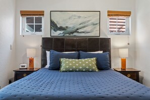Ein Schlafzimmer mit einem großen Bett, zwei Nachttischen, einem Wandbild und Fenstern mit Jalousien.