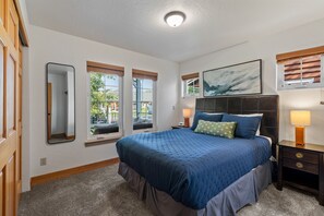 A bedroom with a large bed, a nightstand, a lamp, a painting, and windows with views of the outside. - Serenity at Dover Bay | Sleeps 8 (Sandpoint)