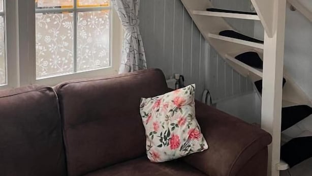 Un sofá marrón con un cojín floral, una escalera blanca y una ventana con cortinas.