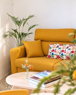 A mustard-colored sofa with floral cushions, a small table with books and a vase of flowers, and a tall green plant beside the sofa. - Maison Sarava – Superior Apartment with Separate Studio & Private Patio (Espira-de-l'Agly)