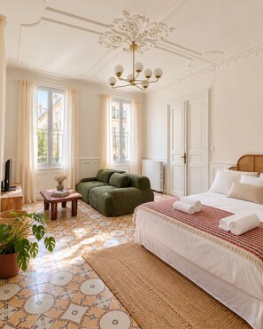 A hotel room with a large bed, a green armchair, a small table with a vase, and a view of buildings through the window. - Maison Sarava – Superior Apartment with Separate Studio & Private Patio (Espira-de-l'Agly)