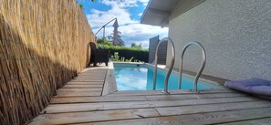 Ein Swimmingpool mit Holzterrasse und Metallleitern, umgeben von einem Bambuszaun und einem Haus.