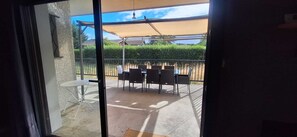 Blick durch eine Glastür auf eine Terrase im Freien mit Tisch und Stühlen, einer Pergola und einem Hecken.