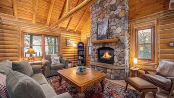 A log cabin interior with a stone fireplace, wooden furniture, and large windows.