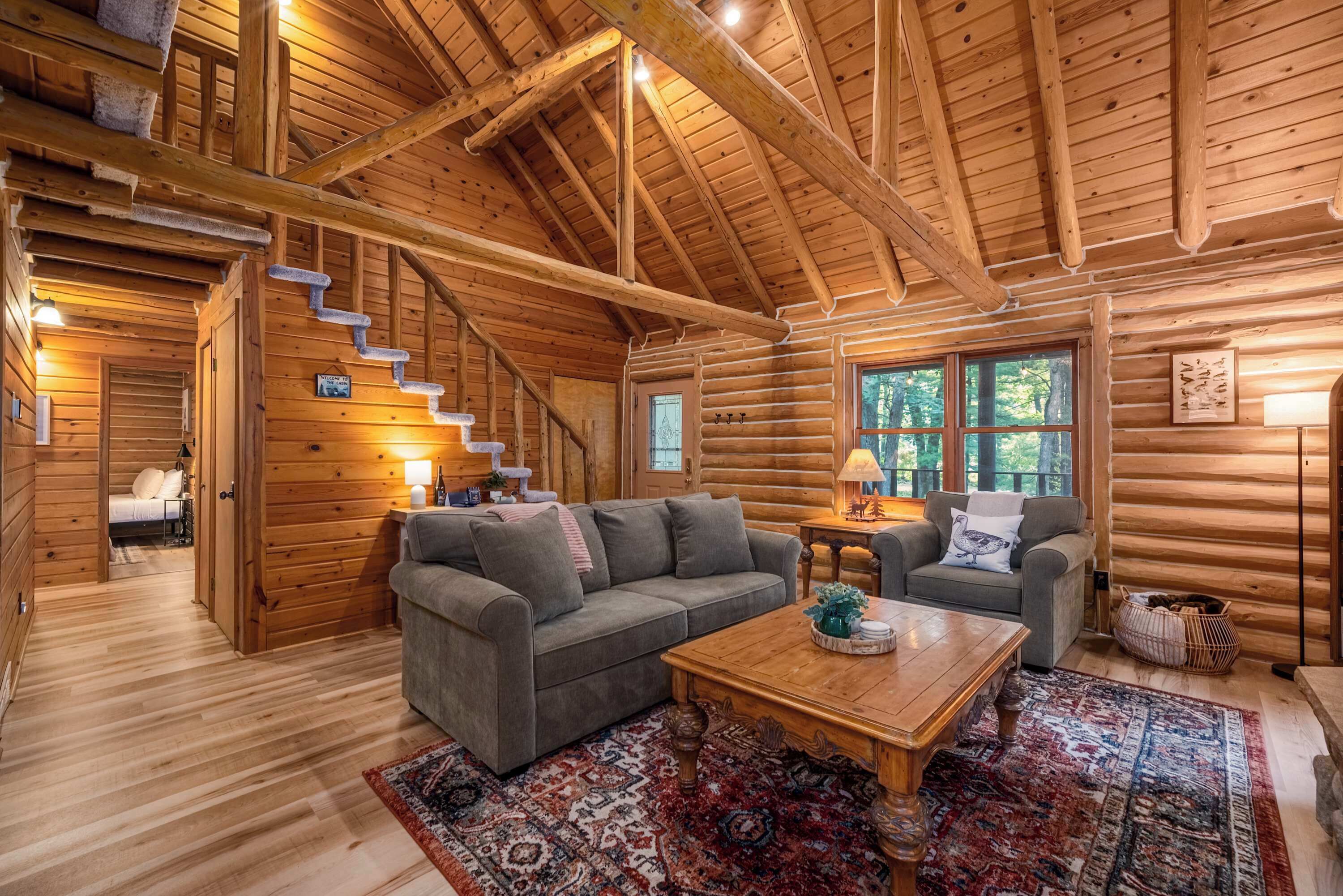 L’intérieur d’une cabane en rondins, avec un canapé, une table basse, un tapis, un escalier et une fenêtre donnant sur la forêt.