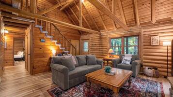 L’intérieur d’une cabane en rondins, avec un canapé, une table basse, un tapis, un escalier et une fenêtre donnant sur la forêt.