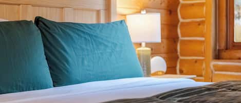 A wooden cabin interior with a bed, pillows, a nightstand, and a window.