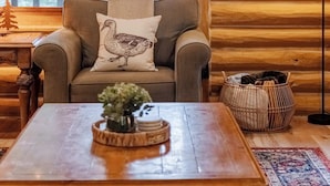 A cozy cabin interior with a wooden log wall, a sofa with a bird-themed pillow, a coffee table with a vase, and a framed picture of birds.