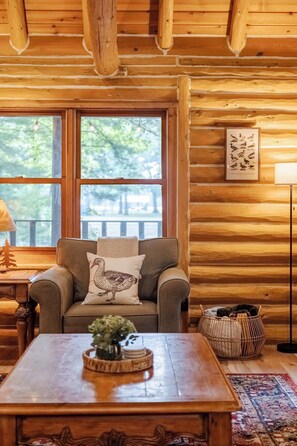 Interior acogedor de una cabaña, con una pared de madera, un sofá con un cojín con motivos de pájaros, una mesa de centro con un florero y un cuadro con aves.