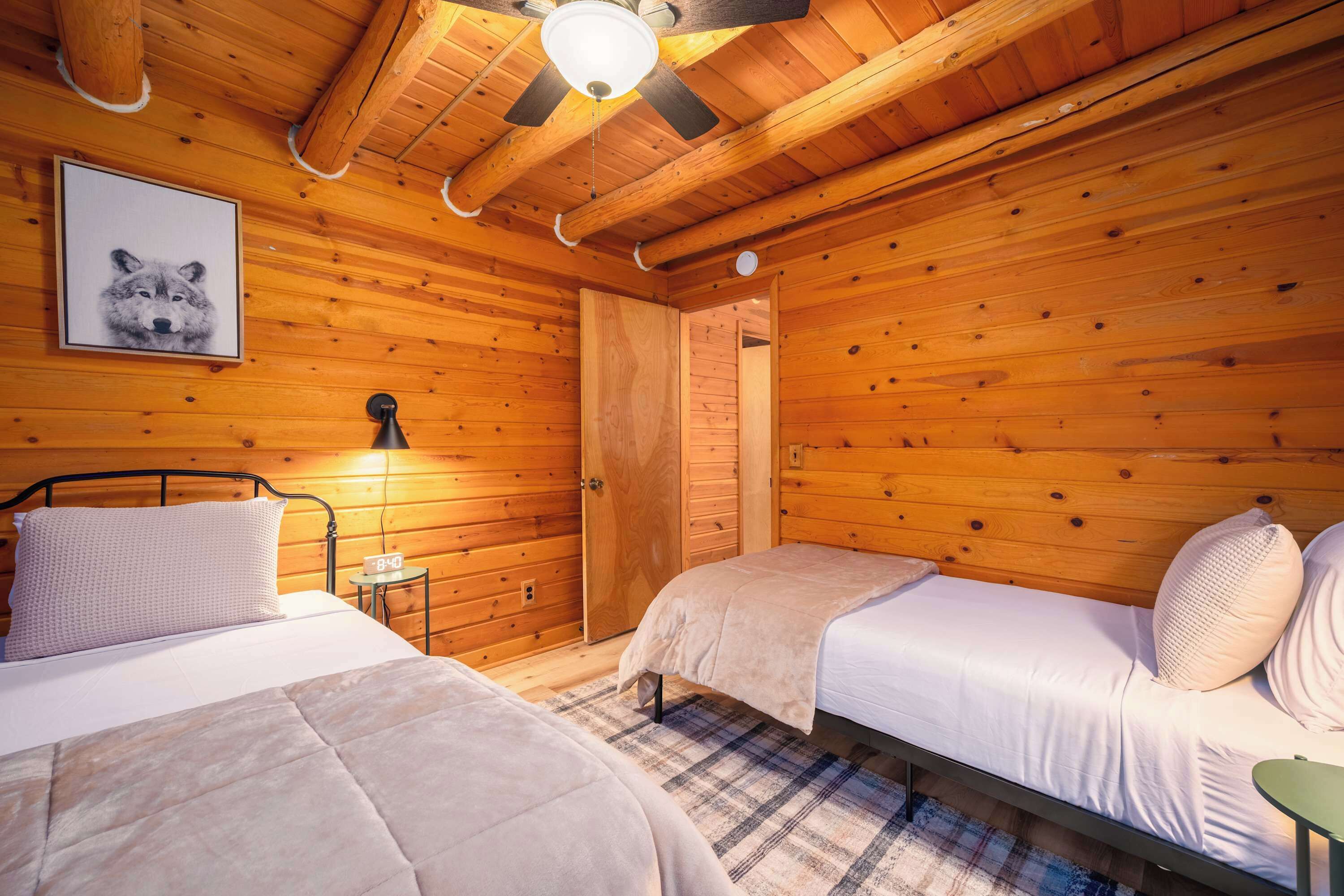 Ein gemütliches Blockhaus-Zimmer mit zwei Betten, einem Deckenventilator, einem gerahmten Bild an der Wand und einem kleinen Tisch mit einer Uhr.