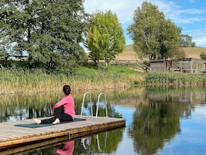A person sitting on a dock by a calm body of water, surrounded by trees and a wooden structure in the background.