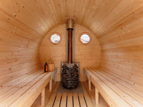 A sauna room with wooden walls, two benches, a central heater, and two windows.