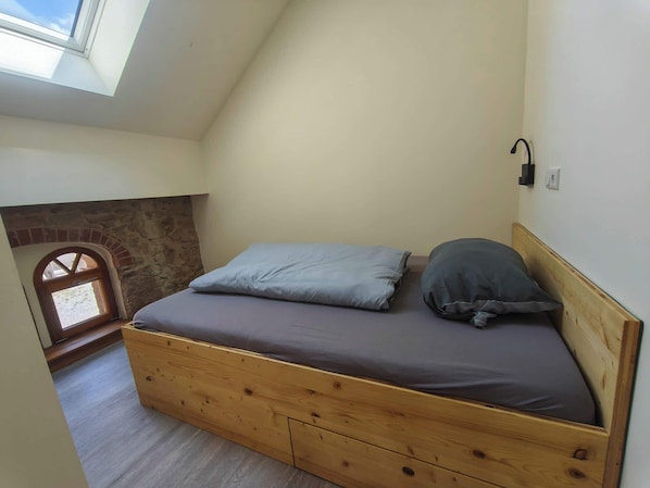 A small, loft-style bedroom with a wooden bed, a stone wall, and a skylight.