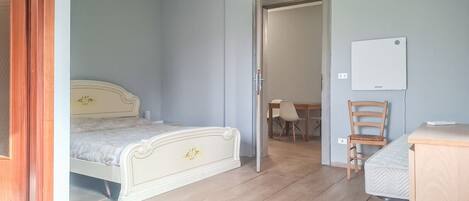 A bedroom with a bed, a desk, a chair, and a door leading to a dining area.