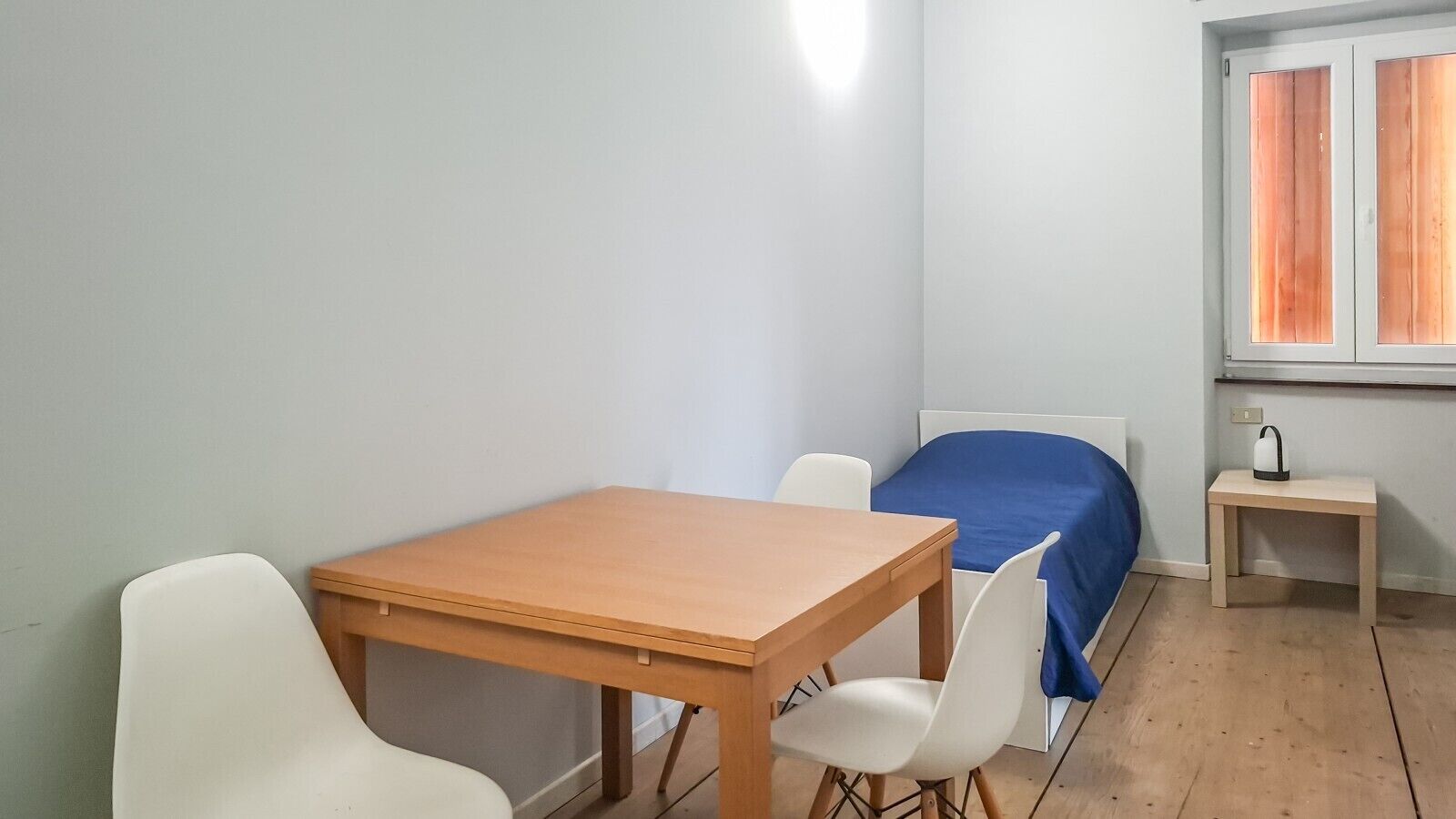 Ein schlichter, kleiner Raum mit einem Holztisch und zwei weißen Stühlen, einem Bett mit blauer Bettdecke sowie einem kleinen Nachttisch mit Wasserkocher.
