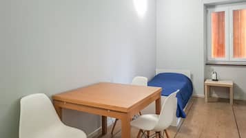 Ein schlichter, kleiner Raum mit einem Holztisch und zwei weißen Stühlen, einem Bett mit blauer Bettdecke sowie einem kleinen Nachttisch mit Wasserkocher.
