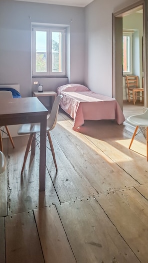 A room with a wooden floor, a bed with a pink bedspread, a table with a chair, and a window.
