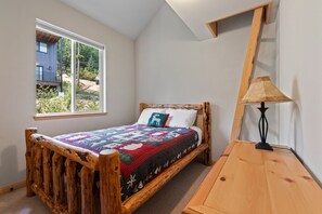Ein Einzelbett mit Holzrahmen, ein Fenster mit Blick auf ein Gebäude und Bäume sowie ein Nachttisch mit Lampe.