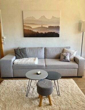 A modern living room with a grey sofa, a round coffee table with candles, a patterned ottoman, and a large landscape painting on the wall. - Charming 1-bedroom apartment in Megève, 10' walking of ski shuttle/village (Megève)