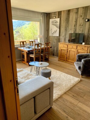 A dining area with a wooden table and chairs, a television on a wooden cabinet, and a view of greenery outside.