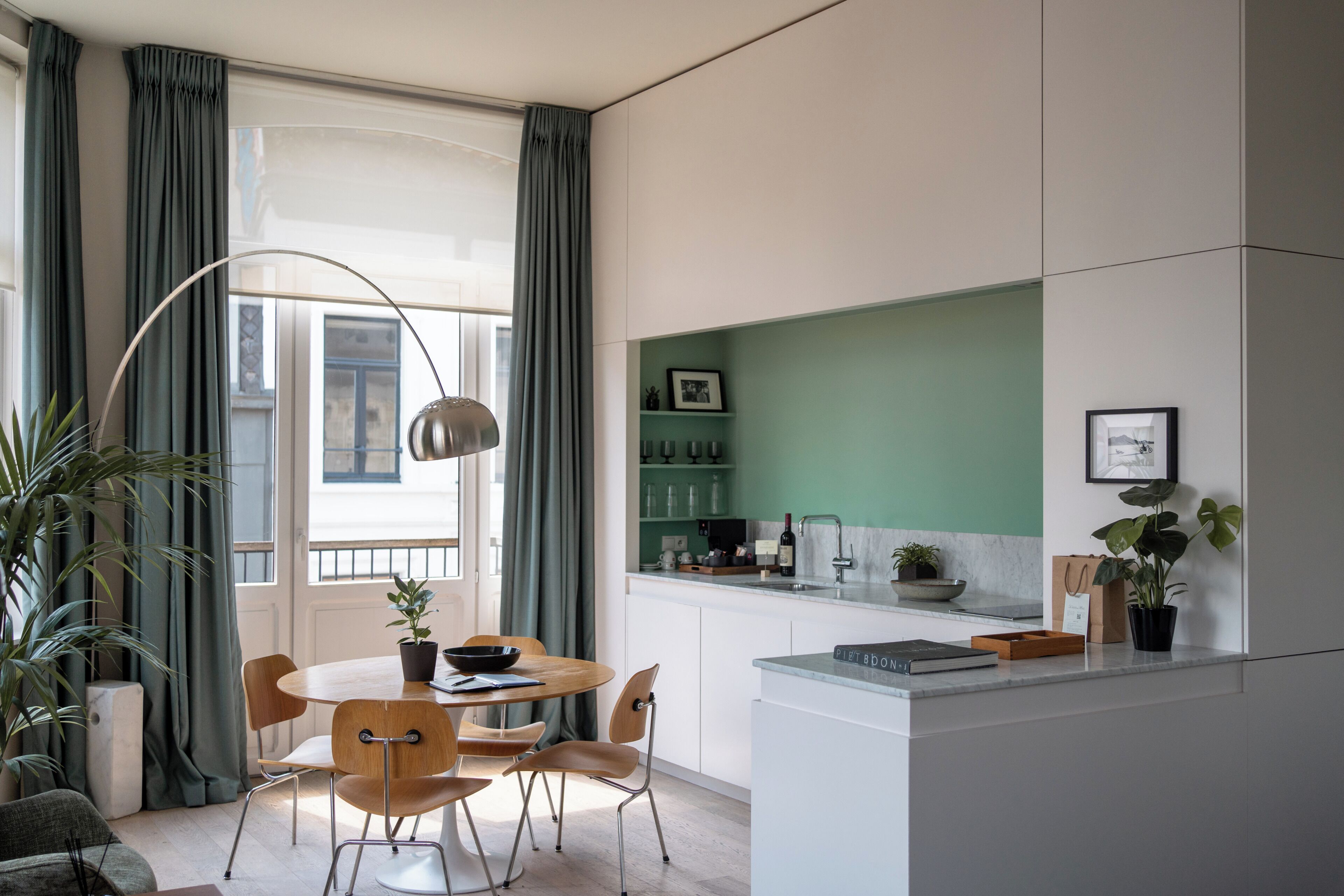 A modern kitchen with a dining area, featuring a round table with chairs, a pendant light, and a view of the city through large windows.