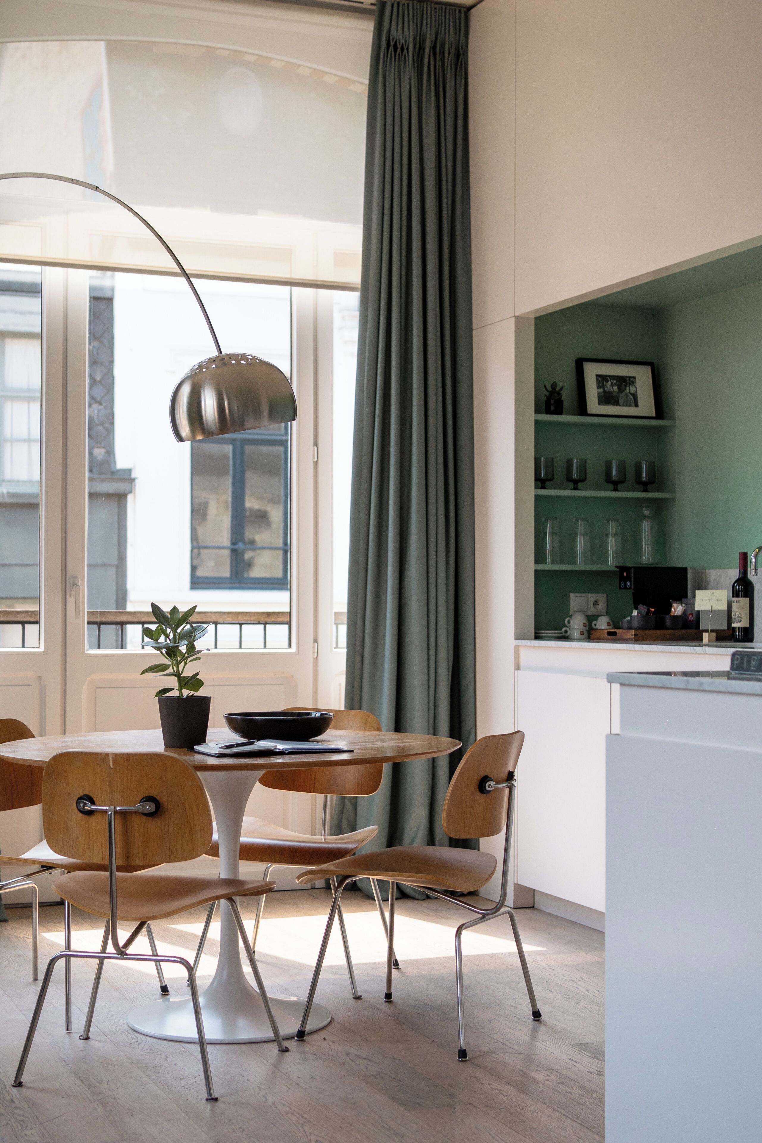 Une cuisine moderne avec une table à manger, des chaises, un luminaire suspendu, une plante en pot et une vue sur la ville grâce à de grandes fenêtres.