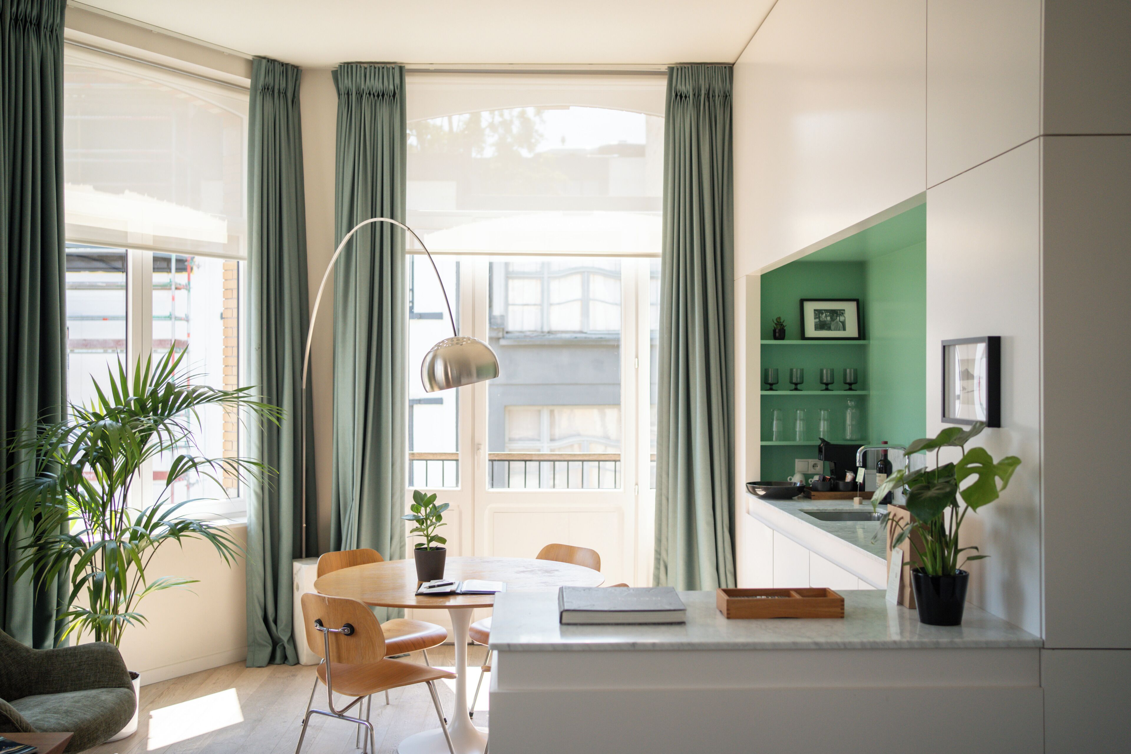 A modern living room with a dining area, large windows with curtains, a floor lamp, and a kitchen with green walls and a sink.