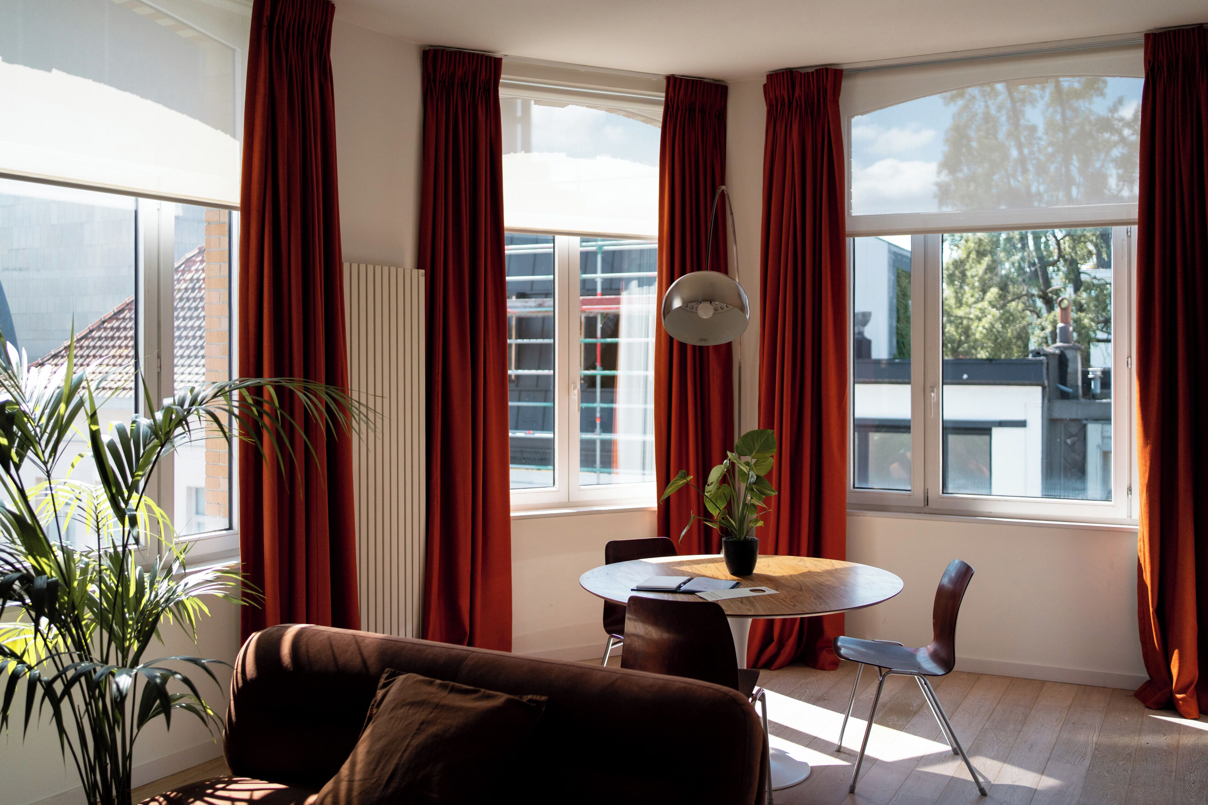 Ein modernes Wohnzimmer mit einem braunen Sofa, einem Esstisch mit Stühlen, einer Topfpflanze und großen Fenstern mit roten Vorhängen.
