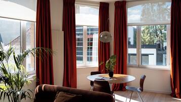 Ein modernes Wohnzimmer mit einem braunen Sofa, einem Esstisch mit Stühlen, einer Topfpflanze und großen Fenstern mit roten Vorhängen.