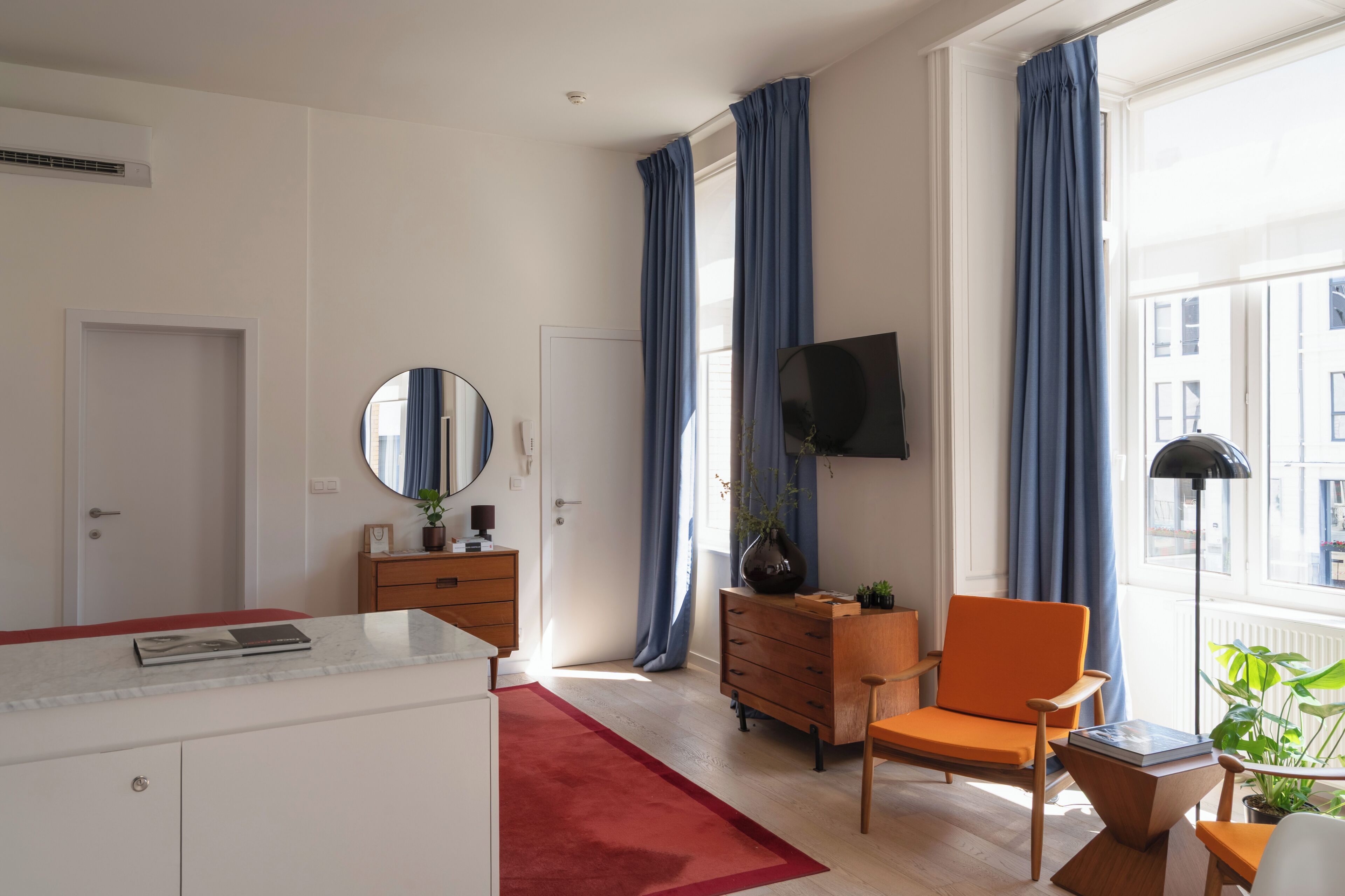 Habitación de hotel moderna con alfombra roja, muebles de madera, un televisor de pantalla plana y ventanas grandes con cortinas azules.