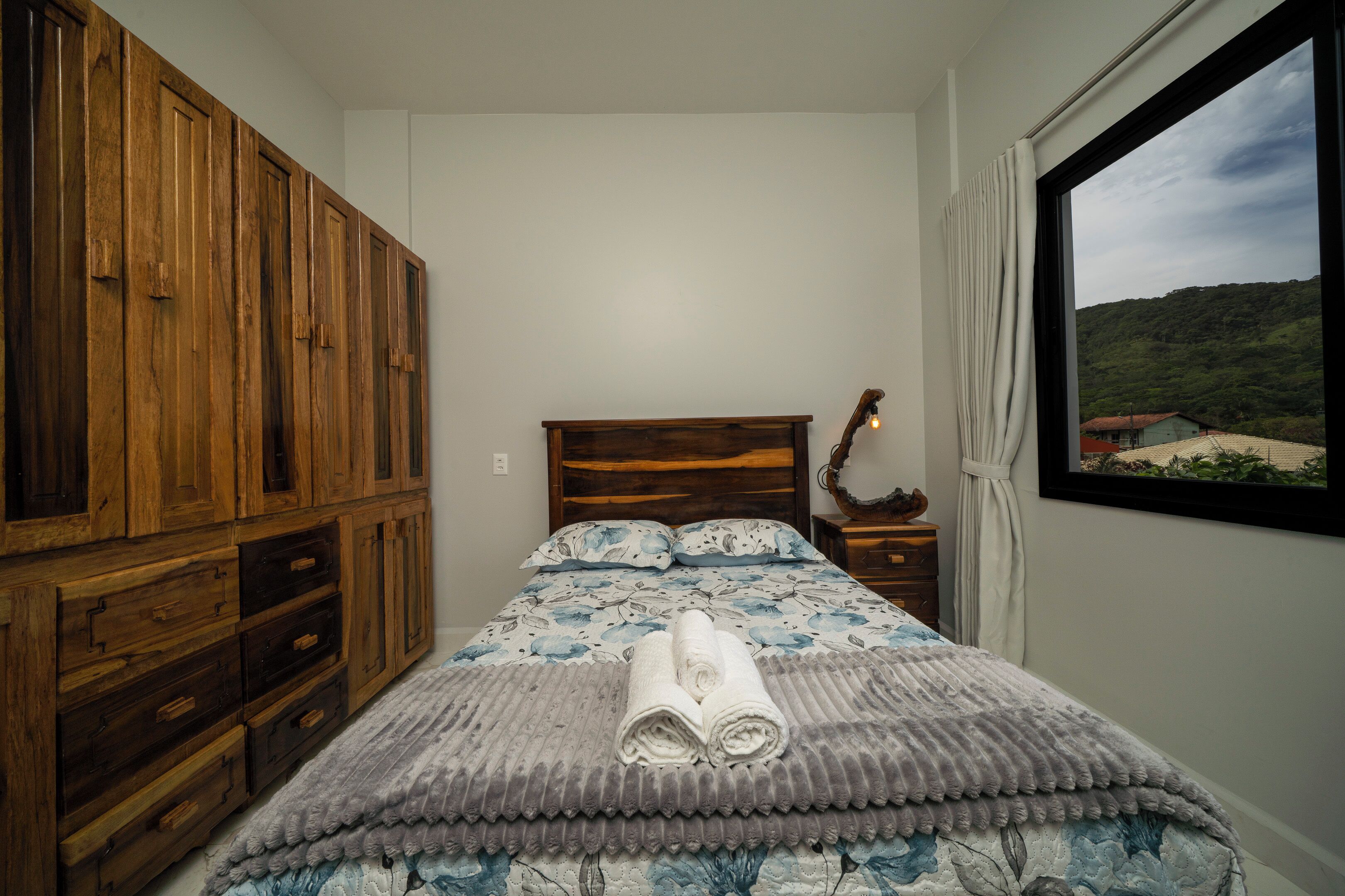 A bedroom with a wooden wardrobe, a bed with a floral duvet, a bedside table with a lamp, and a window with a view of a green landscape.