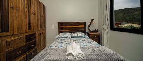 A bedroom with a wooden wardrobe, a bed with a floral duvet, a bedside table with a lamp, and a window with a view of a green landscape.