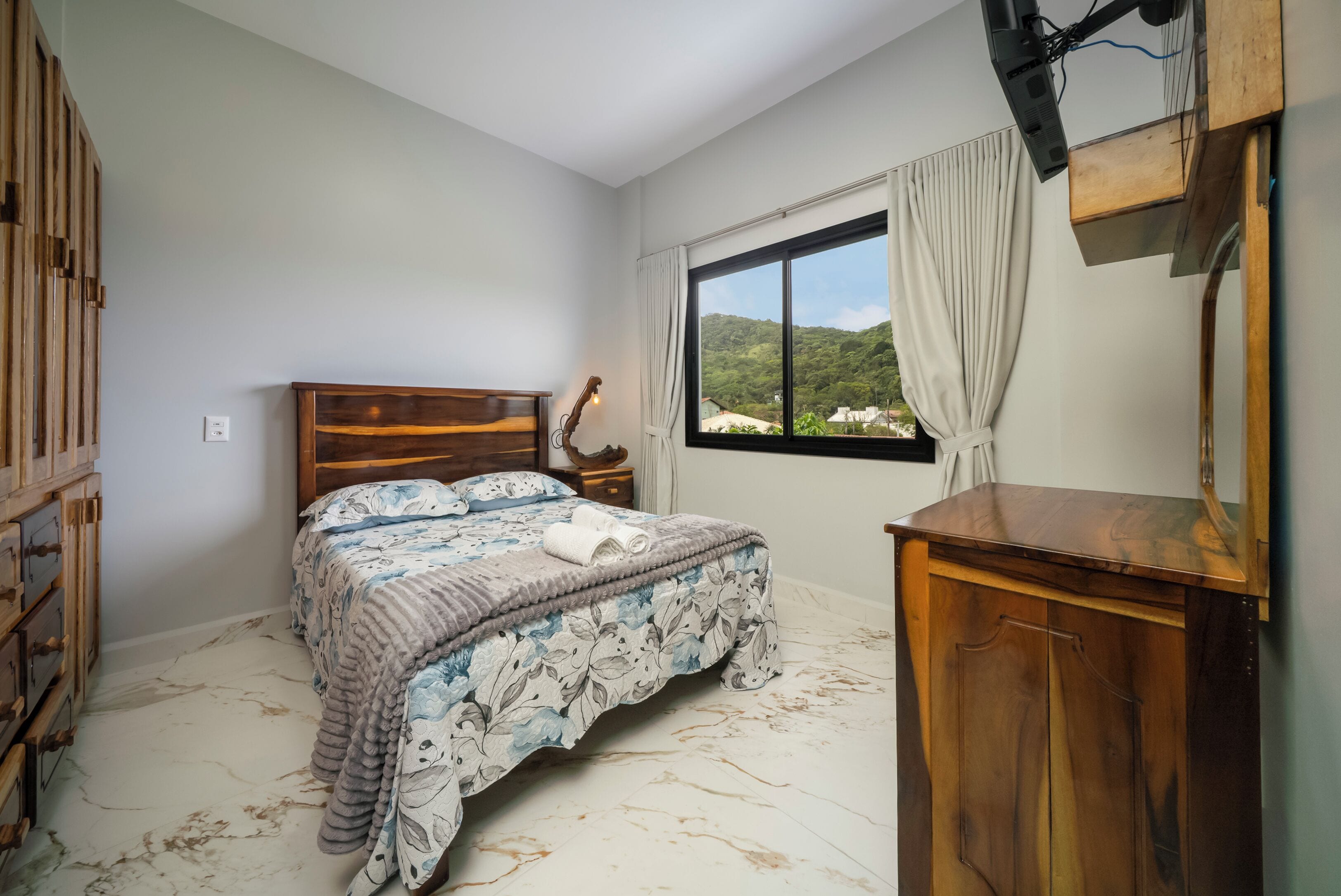 A bedroom with a bed, a wooden nightstand, a dresser, a window with a view of greenery, and a ceiling fan.