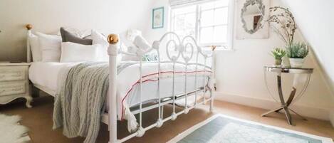 A bedroom with a white metal bed frame, a bedside table, a lamp, a window with a view, and a small table with a plant.